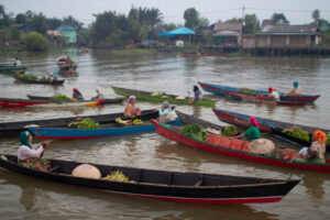Banjarmasin A candid editorial photograph capturing the centuries-old Lok Baintan Floating Market in South Kalimantan, Indonesia, at fajar. Banjarese women vendors, in colorful hijabs and traditional attire, navigate their wooden jukung canoes on the Martapura River. Their boats are a vibrant tapestry, laden with fresh local produce like bananas, leafy greens, and citrus fruits in traditional woven baskets (caping). In a powerful display of ancient tradition, they engage in direct barter trade, exchanging goods rather than monetary currency, their faces conveying a quiet, shared understanding. The backdrop features traditional stilt houses and lush palm trees against a soft, dawn sky. This image documents a rare, authentic, and non-monetary economic system still thriving.