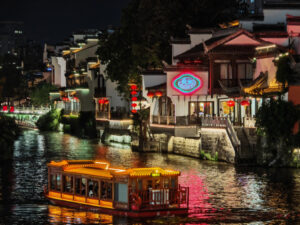 Nanjing A vibrant riverside scene in the historic city of Nanjing, China, featuring a traditional wooden boat illuminated by warm lights and well-preserved buildings with modern neon signs.
