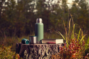 Green thermos, cup and book stand on a stump in the autumn forest.