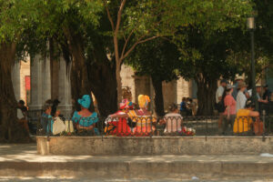 Havana, Cuba 2024 Apr17 - Group of Cuban women dressed in colourful traditional clothing called Rumba dress sitting in a park under trees near a museum in Old Havana. Tourists walking by in park.