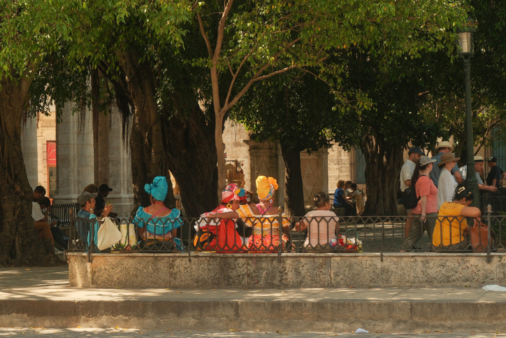 Havana, Cuba 2024 Apr17 - Group of Cuban women dressed in colourful traditional clothing called Rumba dress sitting in a park under trees near a museum in Old Havana. Tourists walking by in park.