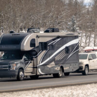 Banff, Alberta, Canada. Apr 4, 2025. A 2023 Thor Magnitude RS36 RV traveling on a road, towing a white pickup truck with items on its roof rack. The surrounding environment appears to be a snowy,