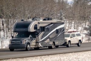 Banff, Alberta, Canada. Apr 4, 2025. A 2023 Thor Magnitude RS36 RV traveling on a road, towing a white pickup truck with items on its roof rack. The surrounding environment appears to be a snowy,