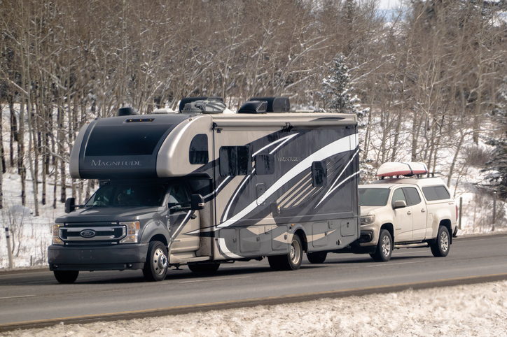 Banff, Alberta, Canada. Apr 4, 2025. A 2023 Thor Magnitude RS36 RV traveling on a road, towing a white pickup truck with items on its roof rack. The surrounding environment appears to be a snowy,
