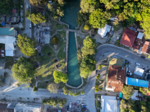Capilay Spring Water Park, San Juan, Siquijor, Philippines – April 14, 2025: Top down aerial view of the spring pool and surrounding roads, buildings, and trees in the town center.