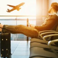 person relaxing in an airport departure lounge, with their feet up on their luggage, watching an airplane take off through a large window