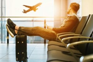 person relaxing in an airport departure lounge, with their feet up on their luggage, watching an airplane take off through a large window